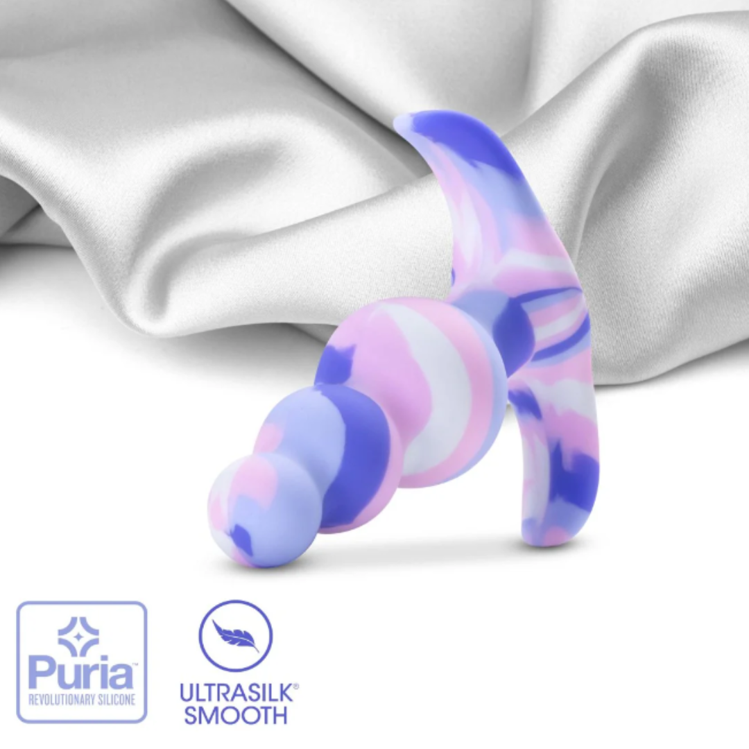 The Avant Twilight Butt Plug resting on a satin fabric background, showcasing its colorful design and highlighting the Puria™ silicone and Ultrasilk® smooth finish branding.