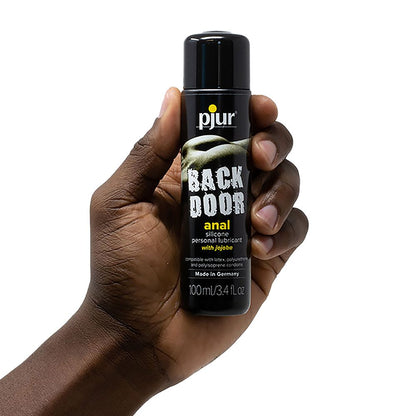 Hand holding a 100ml (3.4 fl. oz.) bottle of Pjur Backdoor Anal Silicone Personal Lubricant with jojoba, suitable for latex, polyurethane, and polyisoprene condoms.